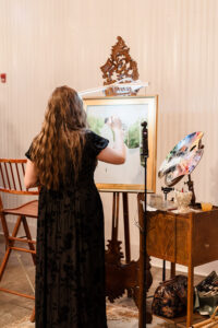 A woman in a black dress paints at an easel, inspired by a Gulf South wedding, with art supplies on a nearby table.