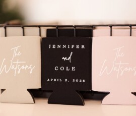 Three can coolers in a basket: two read The Watsons; center says Jennifer and Cole, April 5, 2025. Beige, black, and pink.
