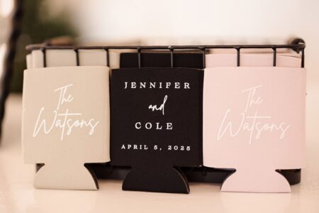 Three can coolers in a basket: two read The Watsons; center says Jennifer and Cole, April 5, 2025. Beige, black, and pink.