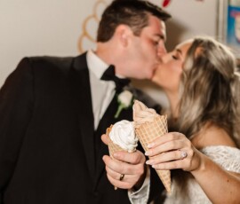 Bride and groom kiss with ice cream cones in hand, faces blurred, red script letters in background—joyful wedding moment.