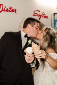 Bride and groom kiss with ice cream cones in hand, faces blurred, red script letters in background—joyful wedding moment.