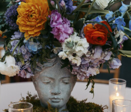 Sculptural centerpiece with colorful flowers arranged in a decorative bust vase surrounded by candles.