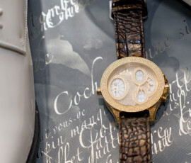 Gold wristwatch with brown leather strap between glossy white shoes on script-covered surface for Gulf South wedding inspiration.