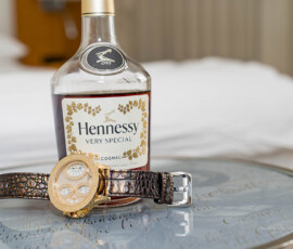 Hennessy Very Special cognac and a gold wristwatch on a glass table before a bed, echoing New Orleans wedding elegance.