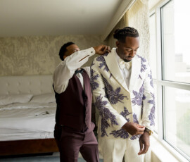 Two men by a bedroom window; one helps the other, who wears a white floral suit—unique Gulf South wedding inspiration.