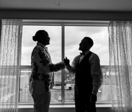 Two men in formal wear shake hands by a sunlit window with city views, exuding New Orleans wedding elegance.