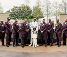 Eleven men in maroon suits, a man in white and purple, and a boy in white pose by a fountain at a Louisiana wedding.