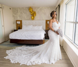 Bride in white lace gown stands by a modern hotel window, bed and gold 50 balloons behind—elegant New Orleans wedding inspiration.