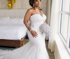 Bride in white lace gown holding jeweled bouquet by window, with gold balloons and a white bed for elegant Gulf South wedding.