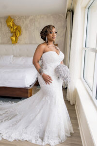 Bride in a fitted lace mermaid gown posing by a hotel window, holding a sparkling bouquet with gold balloons in the background.
