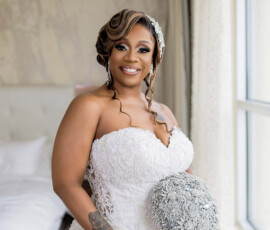 Bride in strapless white lace gown by window, smiling with silver bouquet, drop earrings, and bracelet; classic Gulf South wedding.