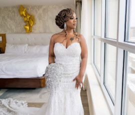 Bride in white strapless gown with jeweled bouquet stands by window; gold balloons and bed in background, elegant curls.