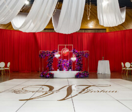 Luxurious wedding venue with red drapes, gold chairs, purple flowers, W monogram floor, stage, and chandelier centerpiece.