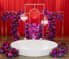 Elegant round white stage with purple, pink, and red flowers, gold frames, chandeliers, and a red backdrop featuring a gold monogram.