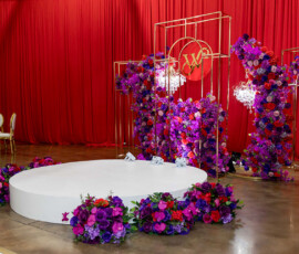 Elegant New Orleans wedding stage with purple and red flowers, gold accents, chandeliers, and red curtain backdrop.