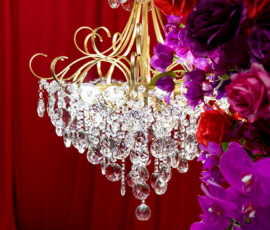 Gold chandelier with crystal drops beside purple, red, and pink flowers on a rich red fabric backdrop—wedding inspiration.
