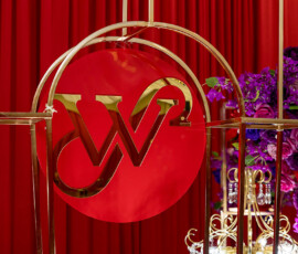 Red circular sign with gold “W2” lettering and frames, set against red curtains and purple florals at a New Orleans wedding.