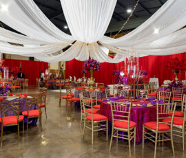 Elegant banquet hall with purple and red tables, gold chairs, tall flowers, draped lights, and dramatic red curtains.