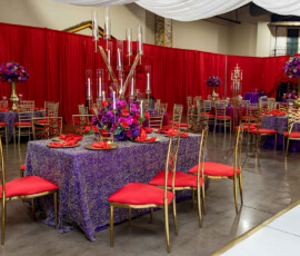 Banquet hall with red and purple decor, gold chairs, floral centerpieces, red curtains, and elegant white ceiling drapes.