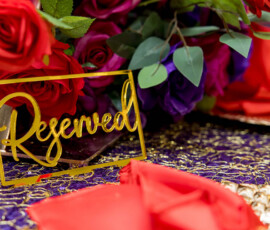 Gold Reserved sign on a table with red roses, purple flowers, greenery, and a shimmering purple-gold tablecloth.