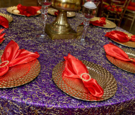 Elegant round table with purple and gold cloth, six gold plates with red napkins, glass candle holders, and gold centerpiece.
