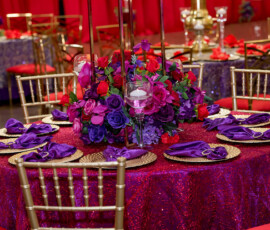 Elegant round table with red sequins, gold chairs, purple napkins, gold plates, and floral candle centerpiece.