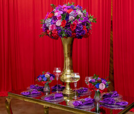 Elegant gold vase with red, purple, and pink flowers on a glass table, wine glasses, and red curtains—New Orleans wedding décor.