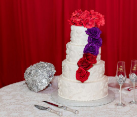 Three-tier white cake with red and purple roses, flanked by champagne flutes and silver bouquet on a white tablecloth, red curtains behind.