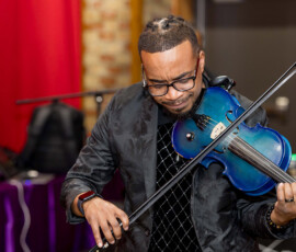 Man in glasses and black jacket plays blue electric violin indoors, red curtain and brick wall behind, evoking New Orleans wedding energy.
