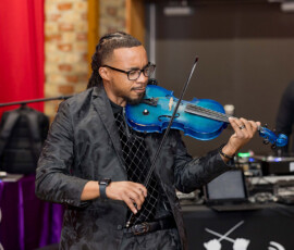 Man in a black suit and glasses plays a blue violin indoors near DJ equipment and a purple-draped table at a Louisiana wedding.