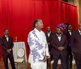 Groom in a white suit with purple patterns and groomsmen in burgundy suits pose by a red curtain at a New Orleans wedding.