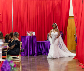 Bride in white gown enters decorated room with red curtains, purple accents, golden chairs, and seated wedding guests.