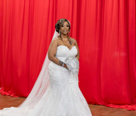 Bride in white lace dress and veil, holding jeweled bouquet and smiling before bright red curtains at New Orleans wedding.