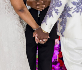 Couple in embroidered white wedding attire hold hands, with officiant and bright flowers in the background. Classic elegance.