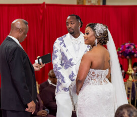 Bride in white lace and groom in floral suit face officiant at wedding with red curtains and guests behind them.