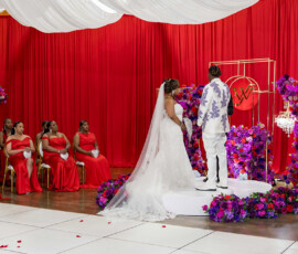 Bride and groom on decorated wedding stage, bridesmaids in red, surrounded by vibrant purple and red flowers—Gulf South style.