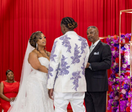 Bride and groom hold hands at a New Orleans wedding with red drapes, purple flowers, and bridesmaids in red dresses.