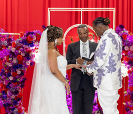 Bride and groom exchange rings in Louisiana wedding, surrounded by purple and red flowers against a red backdrop.