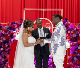 Bride and groom exchange rings with officiant behind them, set against a red backdrop with purple and red floral arrangements.