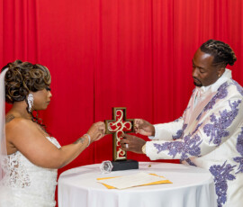 Bride and groom assemble a unity cross at their Louisiana wedding, standing by a round table with documents and a red backdrop.