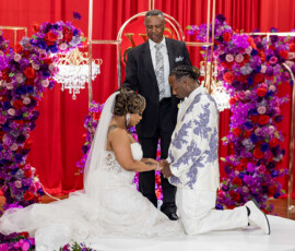 Bride and groom kneel and hold hands before an officiant amid vibrant flowers and red drapes at a romantic New Orleans wedding.