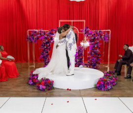 Bride and groom kiss on a round platform with red and purple flowers, flanked by bridesmaids and groomsmen, red curtain behind.