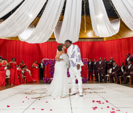 Bride and groom kiss on a white dance floor with red-clad wedding party, draped ceiling, and rose petals in Louisiana.