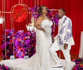 Bride in white gown and groom in white suit with purple designs pose on a red-curtained, flower-filled New Orleans wedding stage.
