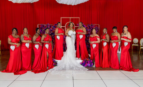 Bride posing with bridesmaids in matching red gowns against a dramatic red and purple floral backdrop.