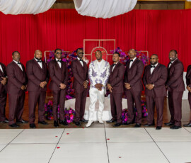 Twelve men stand in a row; eleven wear dark suits, one in white with purple. Red curtains and purple flowers set a wedding scene.