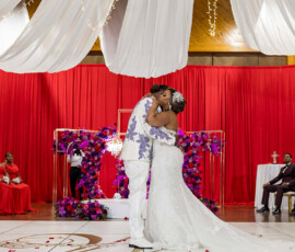 Bride and groom kiss on a decorated stage at their Gulf South wedding, with bridesmaids in red and groomsmen in burgundy.