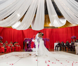 Bride and groom kiss on a red-draped dance floor with wedding party, string lights, and rose petals at a romantic New Orleans wedding.