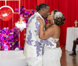 Bride and groom kiss on the dance floor in white with purple accents, framed by a red and purple floral New Orleans backdrop.