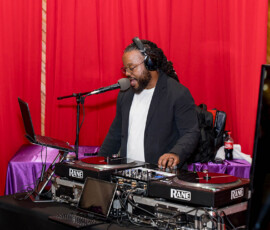 DJ with headphones at turntables and laptop speaks into mic during lively Louisiana wedding; red curtains, purple tablecloth behind.